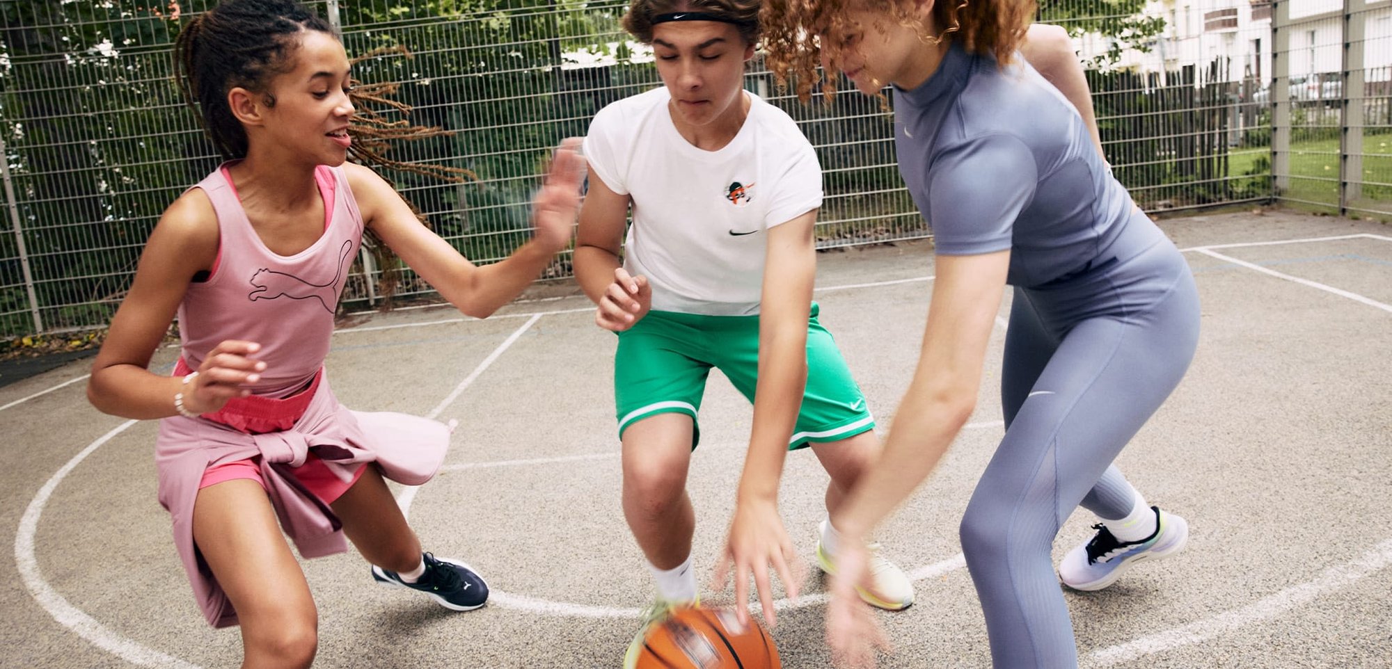 Trois enfants jouent activement au basket-ball sur un terrain. Le premier porte une robe rose de Puma, le second un t-shirt blanc de Nike et un short vert, le troisième un body de sport gris.