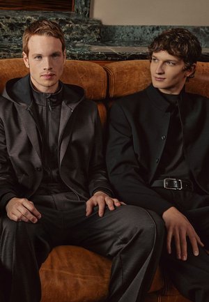 Twee mannen zitten op een bruine leren bank. De een draagt een donkergrijs pak met een capuchonjack; de ander is gekleed in een bijpassende zwarte outfit.