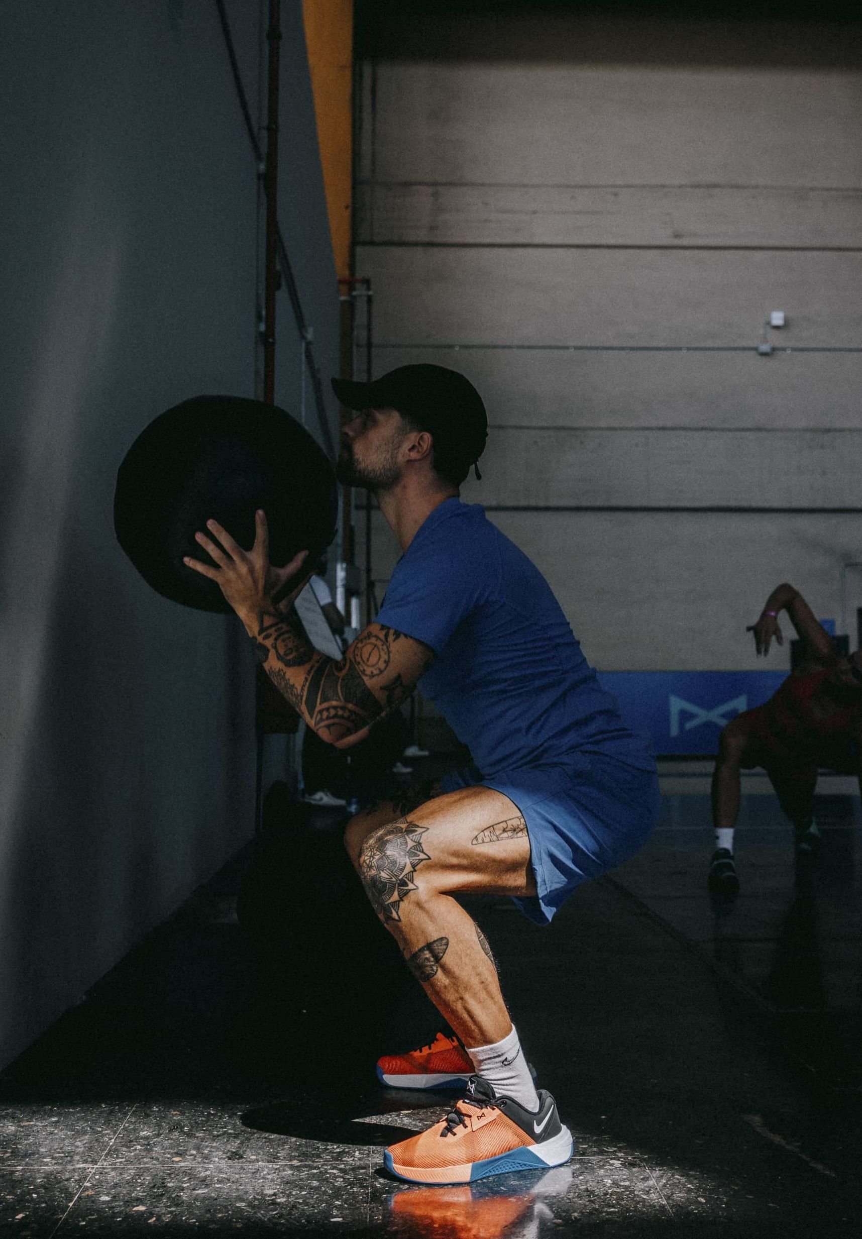 A man in a blue shirt performs a squat while holding a black medicine ball, wearing orange and black athletic shoes. Floor is dark and shiny.