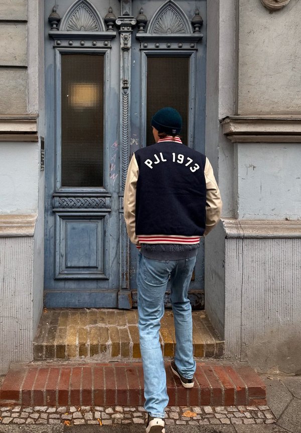 Marineblauw en crèmekleurig varsityjack met rode gestreepte manchetten, met "PJL 1973" op de achterkant. Draagt lichtblauwe jeans en staat voor een decoratieve deur.