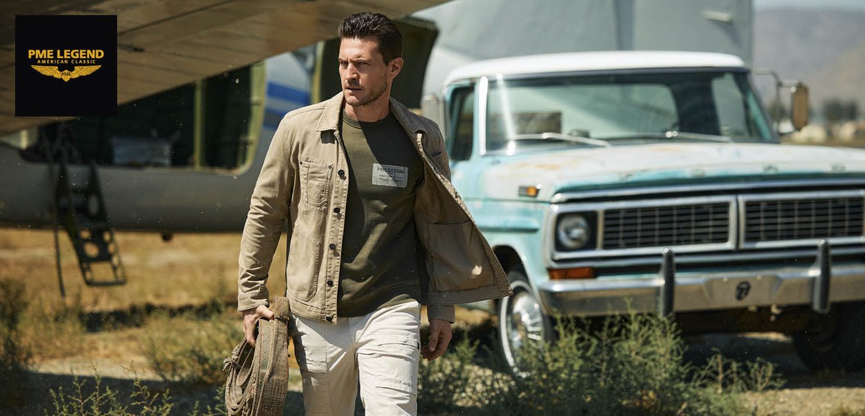 Hombre con chaqueta beige y pantalones blancos camina sosteniendo una cuerda enrollada cerca de un avión antiguo y una camioneta azul vintage en un área de pasto.