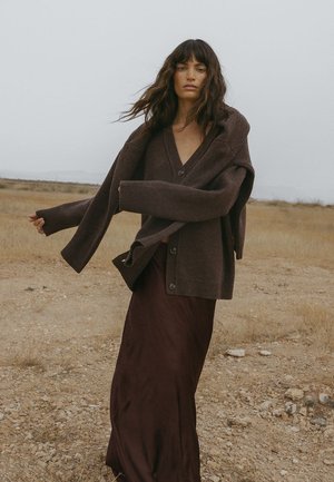 Braun gestrickte Strickjacke mit tiefem V-Ausschnitt, übergroßen Ärmeln und Knopfverschlüssen, kombiniert mit einem langen burgunderfarbenen Rock auf einer braunen Landschaft.