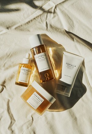 Four skincare products on a textured light fabric: a clear toner bottle, an amber ampoule, a white tube, and a jar with a white lid.