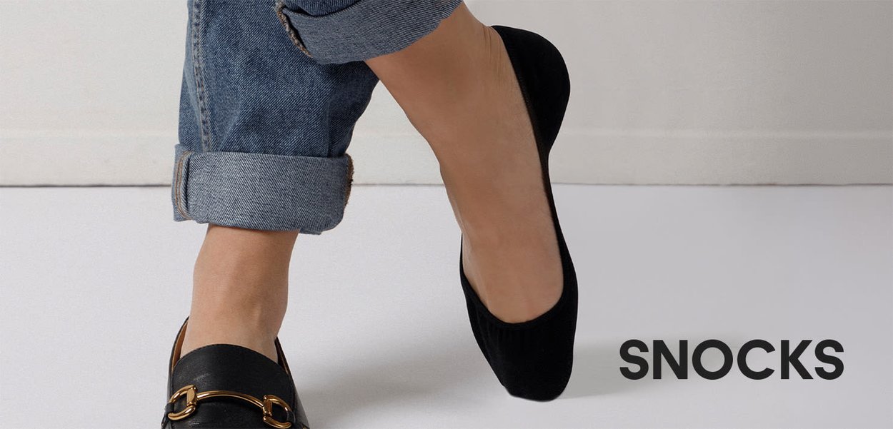 Person trägt hochgekrempelte Jeans und einen schwarzen flachen Schuh mit goldener Schnalle, der andere Fuß ist barfuß mit einer schwarzen unsichtbaren Socke.