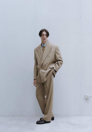 Beige dubbelgeknoopt pak met brede revers en gouden knopen. Lichtblauw overhemd en een gestreepte stropdas. Loszittende broeken en zwarte schoenen.