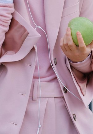 Osoba ubrana w różowy garnitur trzyma w jednej dłoni zielone jabłko, a słuchawki wiszą jej na szyi.