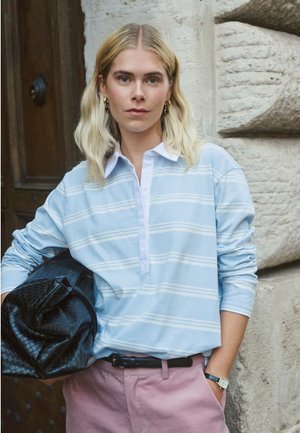 Frau mit blonden Haaren, die ein hellblau gestreiftes Hemd und rosa Hosen trägt, hält eine große schwarze, strukturierte Tasche und steht neben einer Steinmauer.