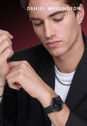 Zegarek z czarnym siateczkowym paskiem, okrągłą tarczą, srebrnymi elementami i czarnym cyferblatem. Noszony na nadgarstku, uzupełniony ciemną marynarką i białą koszulą.