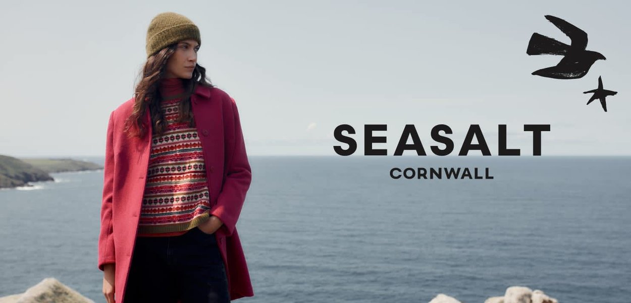 Manteau rouge sur un pull à motifs avec des accents verts, associé à un jean foncé. Le porteur a les cheveux longs et un bonnet vert en tricot, avec en arrière-plan l'océan.