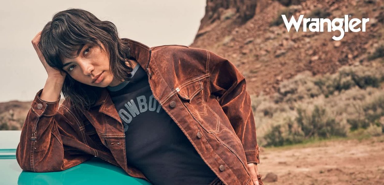 Brun semsket skinnjakke med avslappet passform, med metallknapper og brystlommer, båret over en svart T-skjorte med grafisk motiv. Bakgrunn med steinete terreng.