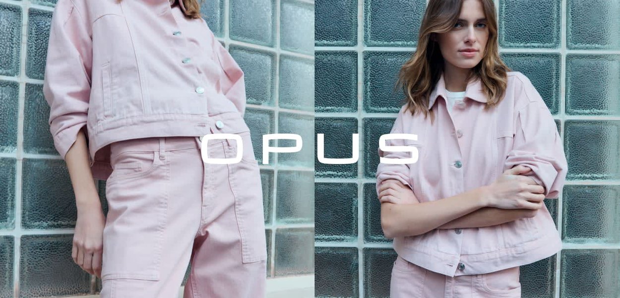 Frau trägt eine hellrosa Jeansjacke und Hose und posiert mit verschränkten Armen und entspannter Haltung vor einer Wand aus strukturierten Glasbausteinen.