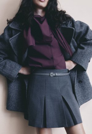Gray pleated skirt with a structured fit, paired with a dark purple shirt and a textured gray coat. Features a buckle belt and button details.