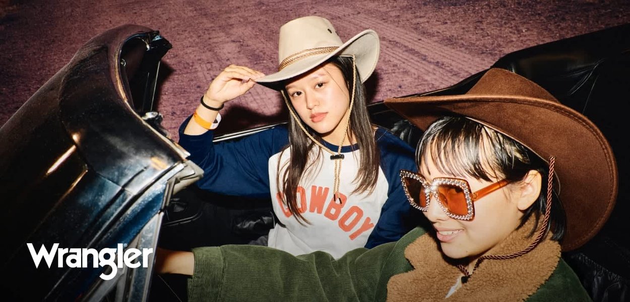 To personer i en svart cabriolet, en iført en beige cowboyhatt og en blå "COWBOY" skjorte, den andre i en brun hatt og store briller.