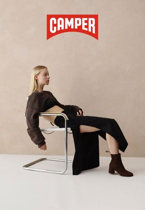 Brauner übergroßer Pullover, schwarzer langer Rock und braune Stiefel. Elegantes Stuhldesign im Hintergrund. Neutrale Wandfarbe. Markenlogo sichtbar.