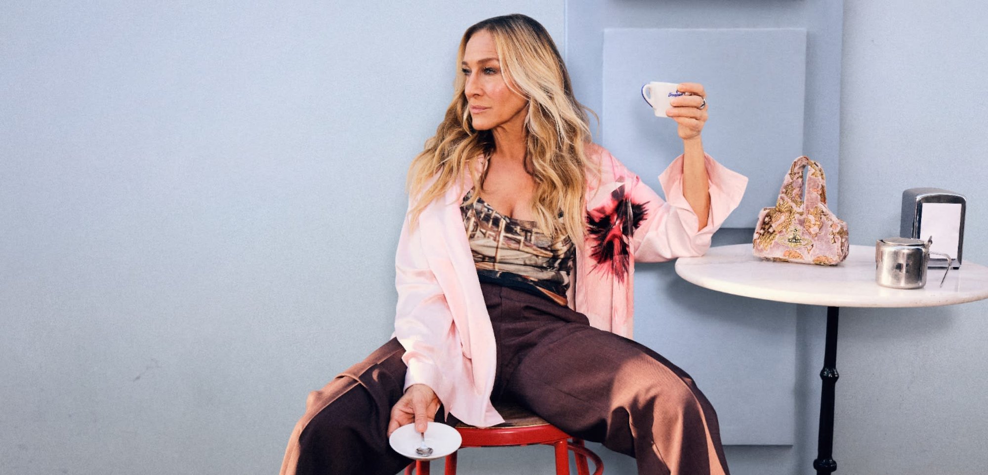 Gaiši rozā pārāk liela izmēra jaka ar ziedu rakstu, virs rakstainas blūzes, brūnas plaša griezuma bikses un rozā papēžu čības, sēžot uz sarkanās krēsla.