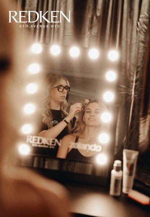 Ein Schminkspiegel mit hellen runden Lichtern, auf dem ein Stylist Haarprodukte auf ein Model anwendet, während die Produkte auf einer dunklen Fläche präsentiert werden.