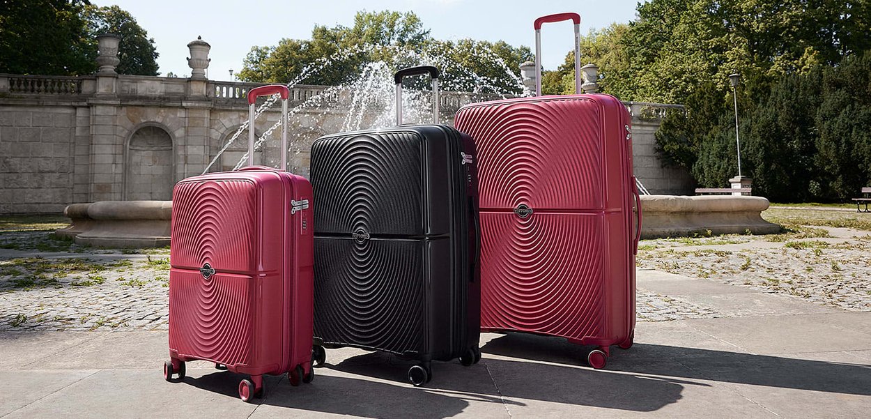 Three hard-shell suitcases in red and black with concentric circle patterns stand on pavement near a fountain in a park.