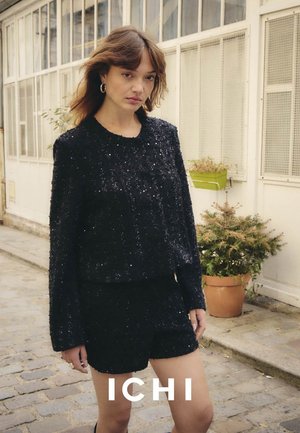 Schwarzes, glitzerndes Zweiteiler-Outfit mit einem texturierten, kurz geschnittenen Oberteil mit langen Ärmeln und taillierten Shorts. Hintergrund zeigt Grünpflanzen.