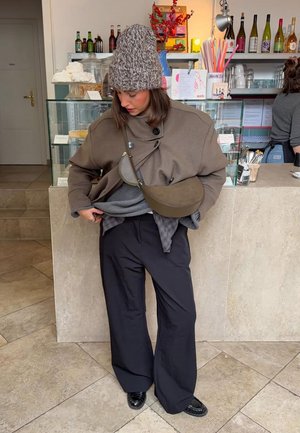 Cappotto marrone oversize con grandi bottoni, maglione grigio, pantaloni neri larghi e berretto lavorato a maglia; borsa a tracolla marrone appoggiata sulla spalla.