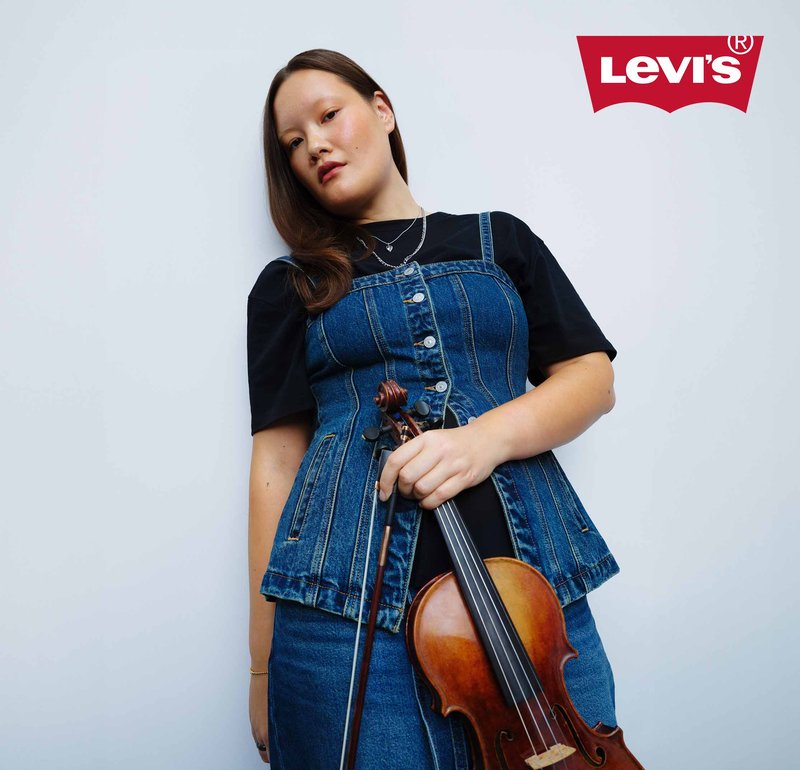 Vrouw in een denim vest en rok die een viool en strijkstok vasthoudt tegen een effen achtergrond, met een Levi's-logo in de hoek.