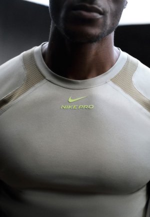 Close-up van een persoon die een beige Nike Pro sportshirt draagt met een neongele Nike-logo en tekst op de borst.
