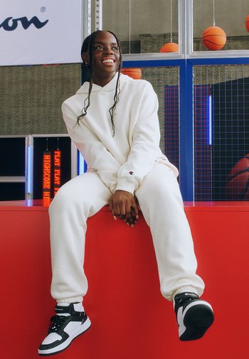 Weißer Kapuzenpullover und Sweatpants in lockerer Passform, kombiniert mit schwarz-weißen Sneakers. Heller Hintergrund mit Basketballs und Neonlichtern.