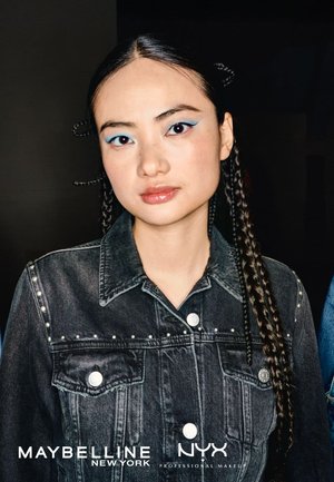 Femme jeune avec de longs cheveux tressés porte une veste en denim noir à clous et un eyeliner bleu audacieux en ailes sur fond sombre.