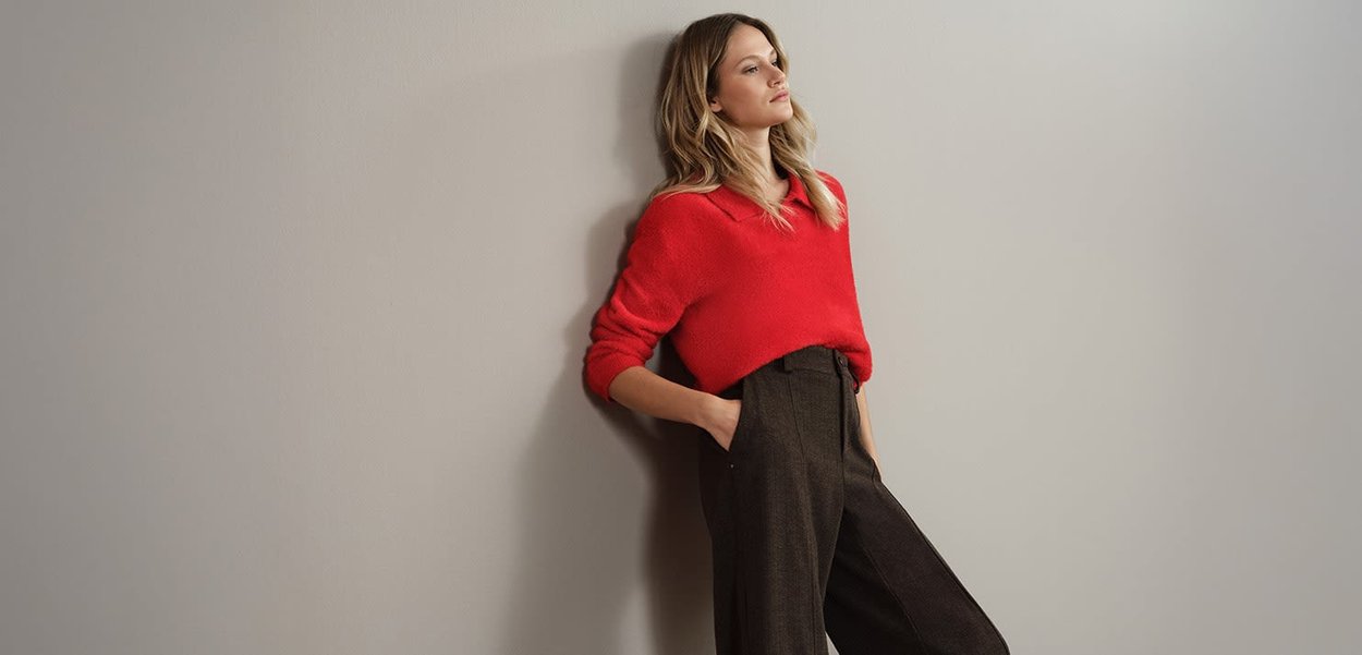 Pull rouge texturé avec col assorti, porté avec un pantalon à carreaux marron taille haute. Le modèle est adossé à un mur clair.