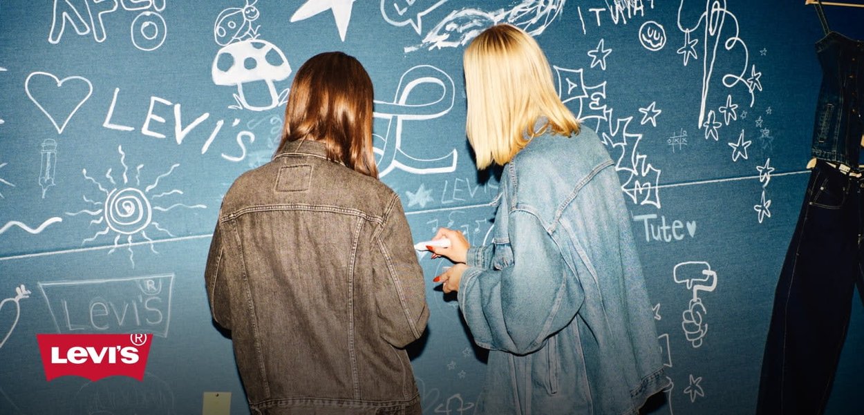 Two individuals wear denim jackets against a blue wall covered in white doodles, including stars and the brand name "Levi's."