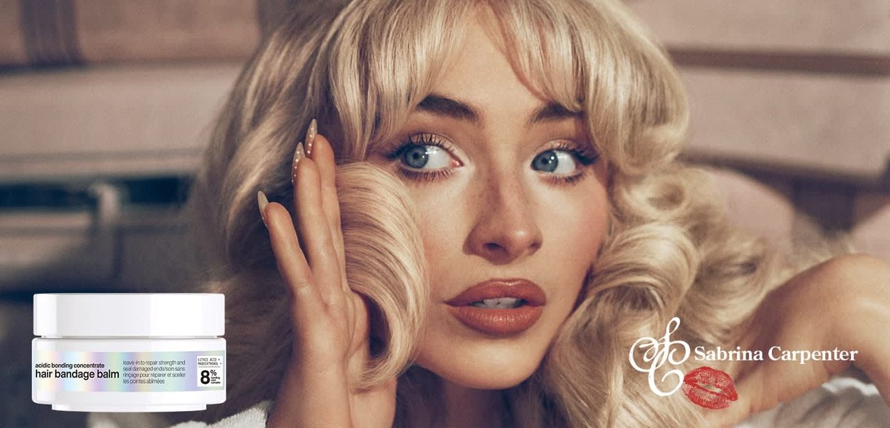 Blond kvinne med krøllete hår som berører ansiktet sitt, og promoterer hårbandasje-balsam vist i en hvit krukke ved siden av henne.