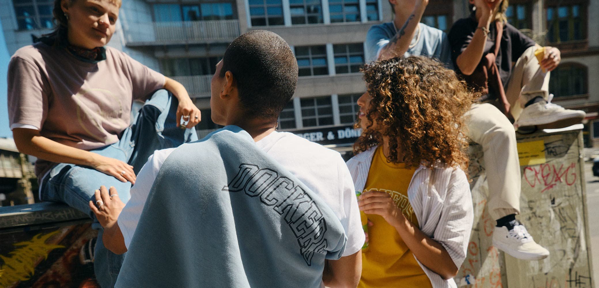 Groupe décontracté dans un environnement urbain ; tenues variées avec des t-shirts graphiques et des vestes légères, y compris une veste bleue avec "DOCKERS" en texte noir.