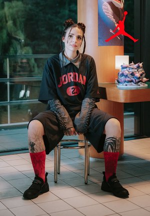 Jersey a rete nera con la scritta "JORDAN" in rosso, maniche lunghe grigie, pantaloni corti neri, calze rosse e scarpe nere con dettagli argentati. Sfondo in legno.