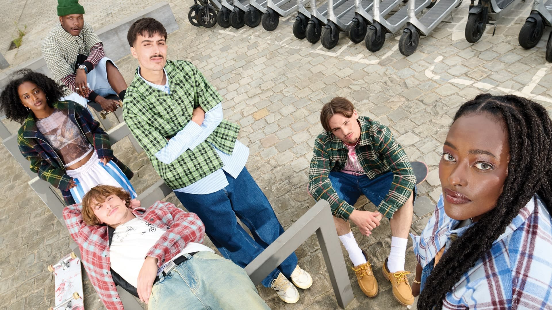 Zes personen zitten in een informele buitenomgeving. Ze dragen uiteenlopende geruite outfits met denim, en een skateboard is zichtbaar voor hen.