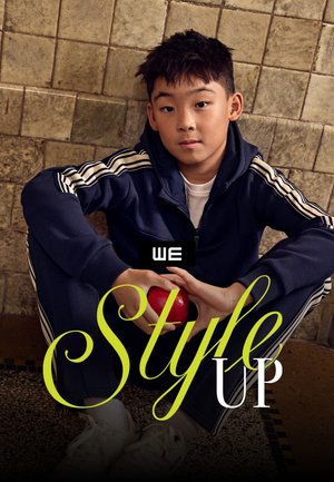 Ragazzo in tuta sportiva blu navy seduto a gambe incrociate contro un muro piastrellato, che tiene una mela rossa con sovrapposto il testo "we style up".