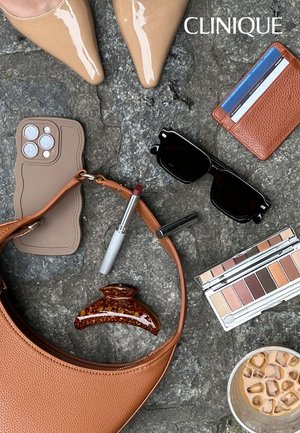 Borsa a mano in pelle marrone, custodia per telefono beige, articoli per il trucco, occhiali da sole scuri, portafoglio marrone e una molletta per capelli su una superficie di pietra. Scarpe con tacco di un marrone chiaro visibili.
