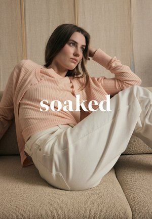 Woman in a light pink sweater and white pants reclining on a beige sofa, looking thoughtful, with the word "soaked" overlaid.