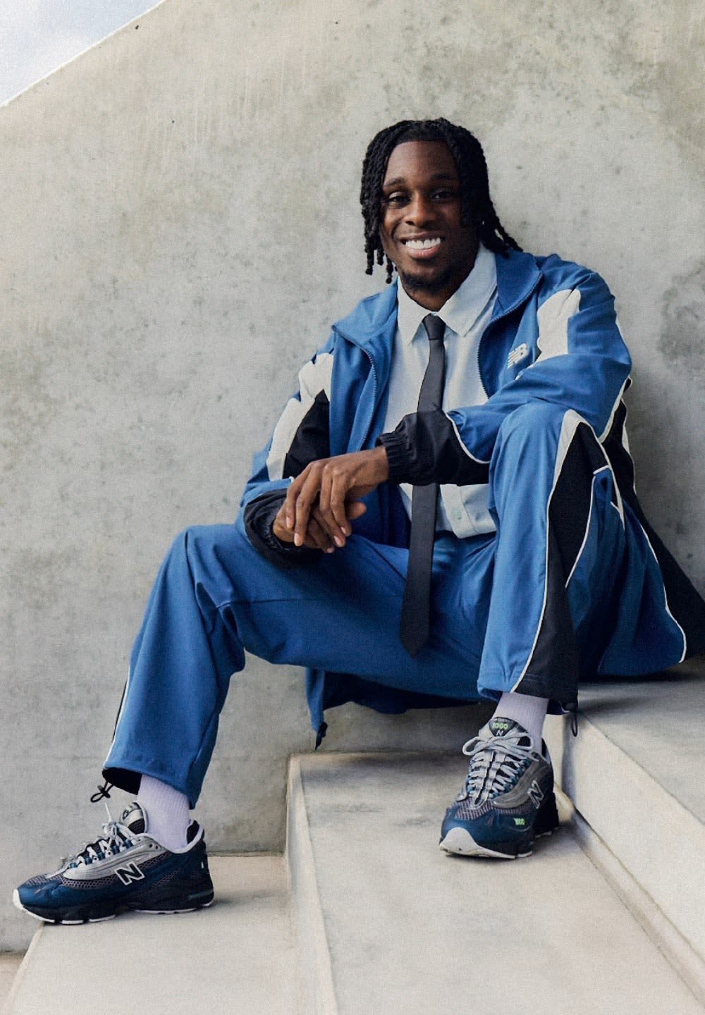 Survêtement de sport bleu avec des accents noirs, porté avec une chemise blanche et une cravate noire. Présente des baskets noires et grises, assis sur des escaliers.