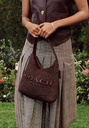 Person trägt eine braune Lederweste und einen karierten Rock und hält eine braune geflochtene Coach New York Handtasche im Freien mit Blumen im Hintergrund.