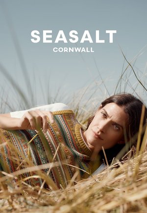 Vrouw liggend in droog gras, gekleed in een gestreepte mouwloze trui over een wit shirt, onder een heldere blauwe lucht met de tekst SEASALT CORNWALL er boven.
