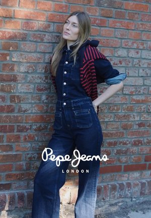 Donkerblauwe denim overall met voorzakken en knopen, gelaagd met een zwart en rood gestreept shirt met lange mouwen. Achtergrond van een baksteenmuur.