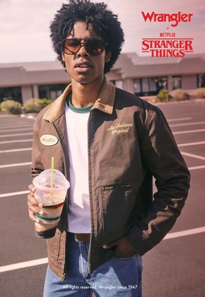 Braune Jacke mit beigem Kragen, grüner Stickerei und einem Aufnäher. Modell trägt Sonnenbrille, hält ein Getränk, kombiniert mit weißem Hemd und blauer Jeans.