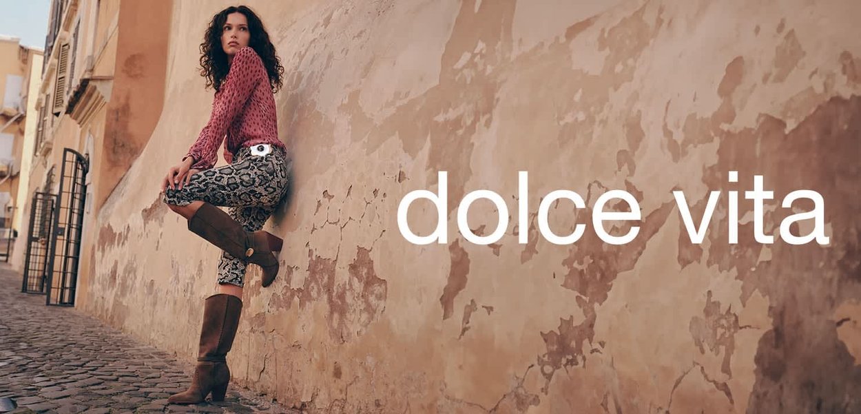 Kniehoge bruine laarzen, gemusterde grijs en zwarte broek, en een roze blouse met lange mouwen tegen een gestructureerde, afbladderende muur. De tekst luidt "dolce vita."