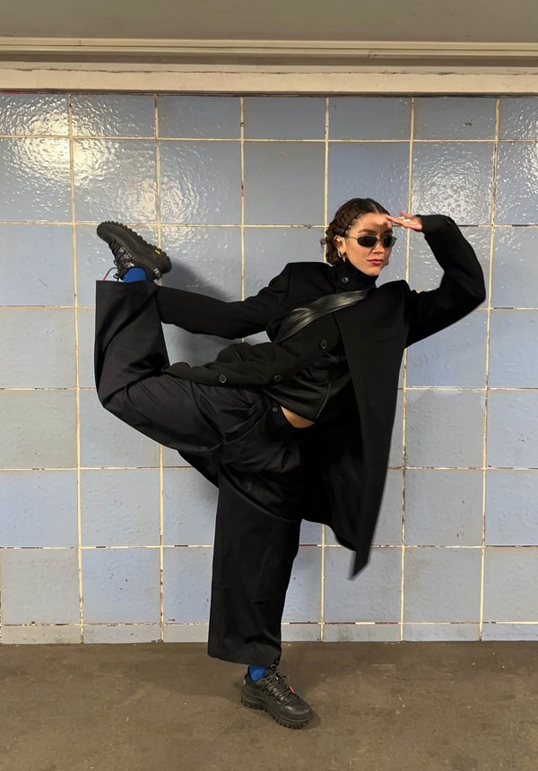 Manteau noir oversize avec une couche intérieure ajustée, pantalon noir baggy, chaussures noires chunky et lunettes de soleil. Devant un mur carrelé bleu clair.