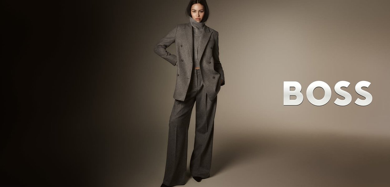 Grijs op maat gemaakt pak met een dubbelgeknoopte blazer en wijde, hooggesneden broekspijpen. Gebreide coltrui eronder. "BOSS"-logo zichtbaar.