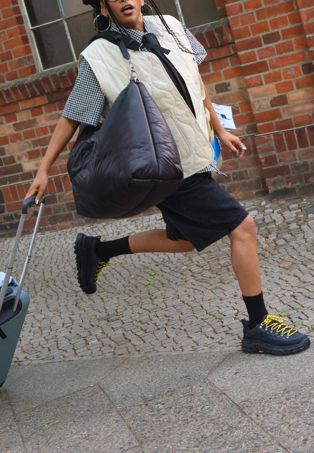 Black puffer bag, beige quilted vest over a checkered shirt, black shorts, and trainers with yellow accents. The person is in motion beside a suitcase.