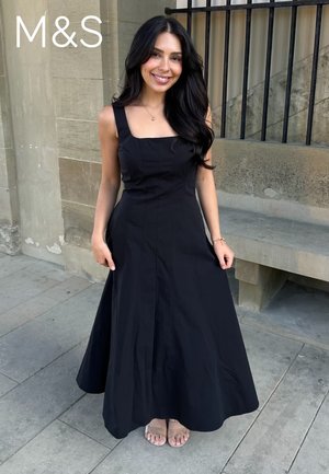 Frau mit langen schwarzen Haaren, die ein ärmelloses schwarzes Maxikleid und Sandalen trägt und draußen vor einer Steinmauer und einer Bank steht.