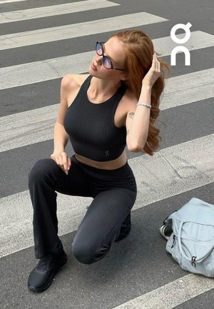Woman with long red hair in black ribbed activewear and sunglasses kneels on a city crosswalk near a light gray backpack.