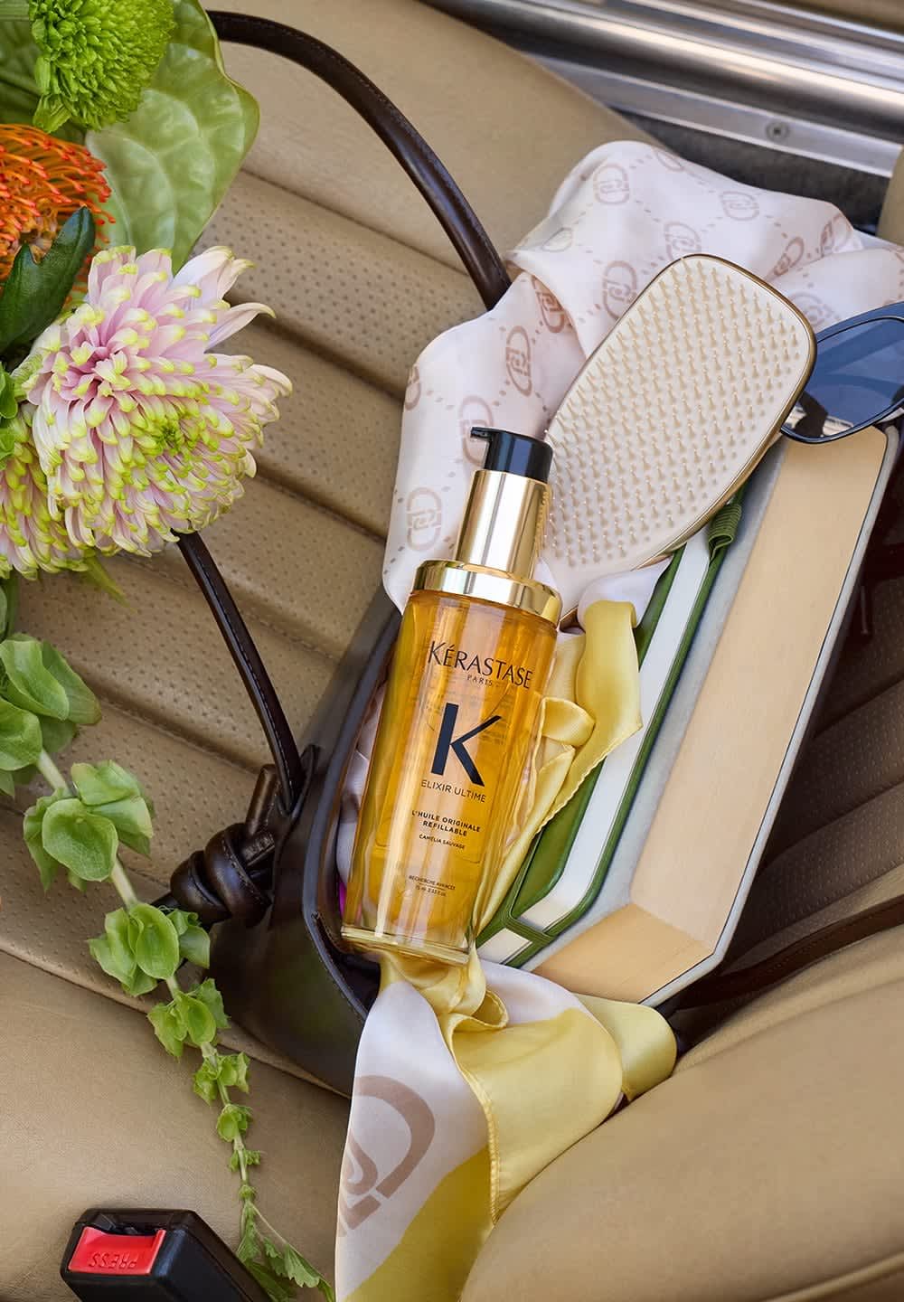 Kérastase Elixir Ultime oil in a gold and transparent bottle, placed inside a brown leather bag with a cream scarf and flowers.