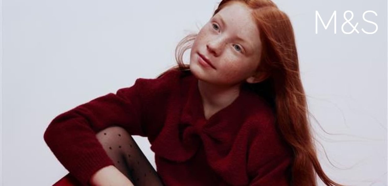 Czerwony dziany sweter z kokardą i długimi rękawami, w połączeniu z czarnymi rajstopami w groszki. Modelka ma piegi i długie rude włosy.