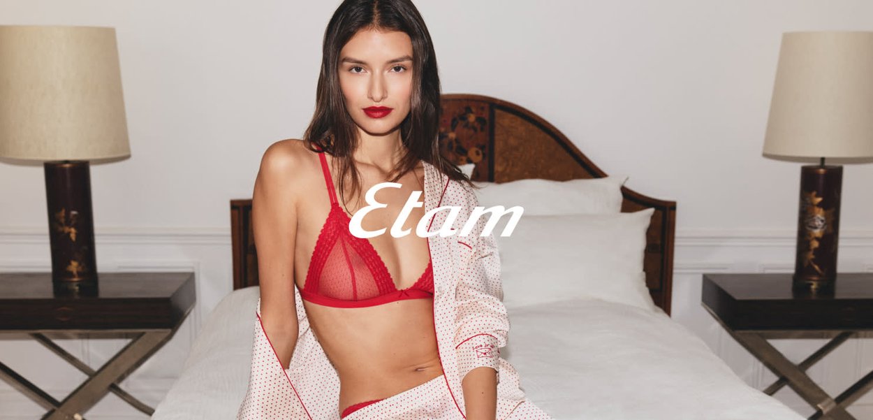 Femme portant une lingerie rouge et une chemise blanche à pois assise sur un lit entre deux tables de chevet avec des lampes.